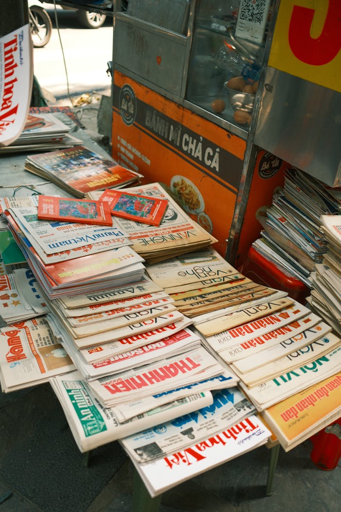 Outdoor vendor table displaying a variety of stacked newspapers in an urban setting.