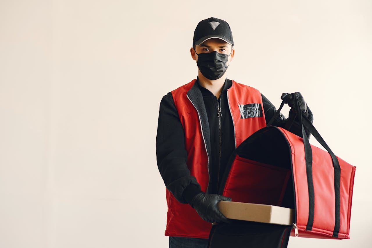 gallery-03 Delivery man wearing a mask and gloves with a red thermal bag and box indoors.