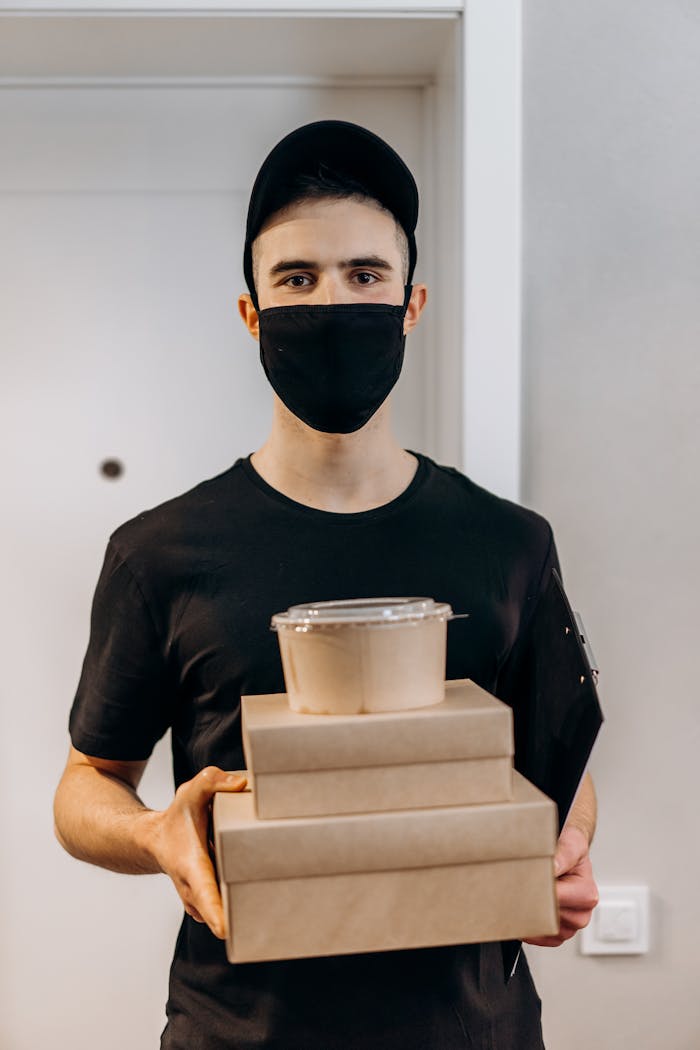 gallery-01 A young delivery man in a black mask holds food boxes indoors, ready for delivery.
