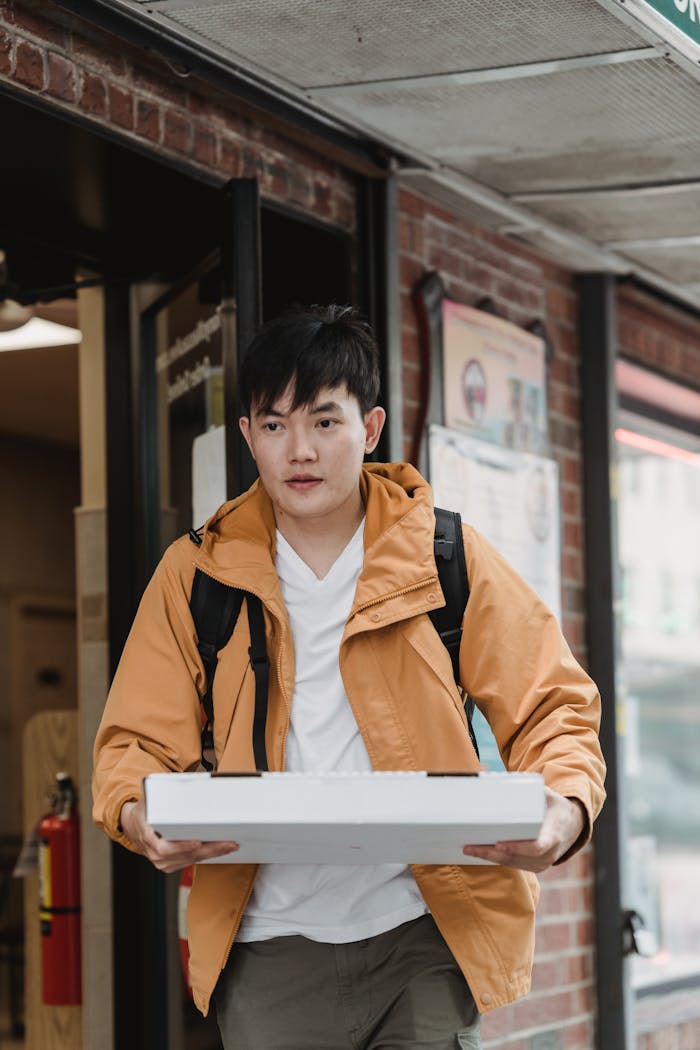 gallery-05 A young man in a yellow jacket holds a pizza box while exiting a building, offering a delivery service.